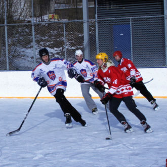 Five ice hockey players skate after the puck on an outdoor ice rink.