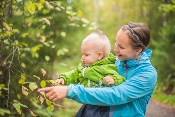 A woman and a baby are pointing at tree leaves beside a forest path.