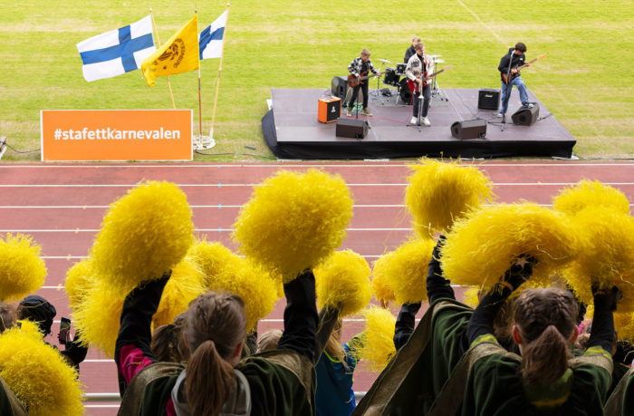 Un groupe de musiciens est en train de se produire sur une scène aménagée devant une piste de course tandis que des pom-pom girls agitent des pompons jaunes.