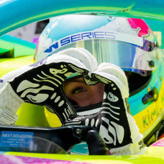 A helmeted racecar driver makes a heart shape with her hands.