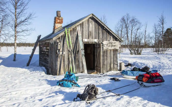Des skis et des luges sont visibles devant une petite bâtisse en bois entourée par un paysage enneigé.