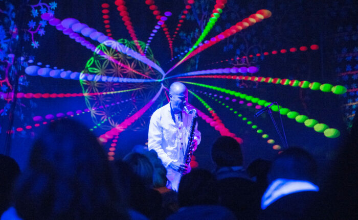 A man in white plays a bass clarinet on a stage in front of colourful light patterns.