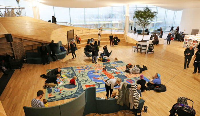 In a large room with a high ceiling, parents and small children lounge and play on sofas and rugs.