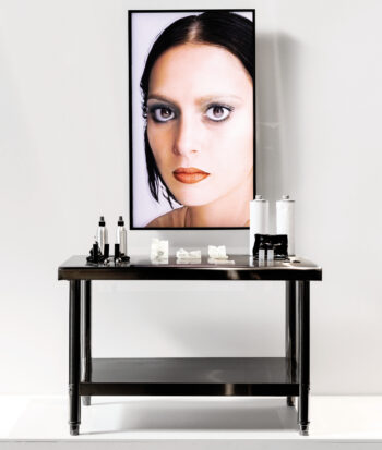 Una mesa de maquillaje con frascos y utensilios y, en lugar del espejo, una pantalla rectangular que muestra el rostro de una mujer.