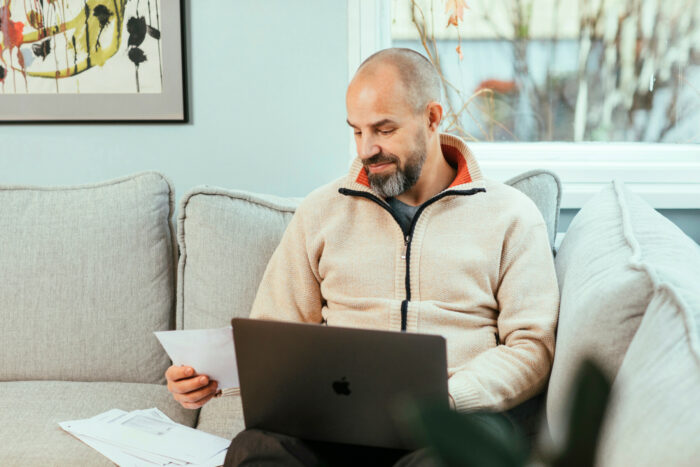 A man sits on a sofa with a piece of paper in one hand and a laptop open in his lap.