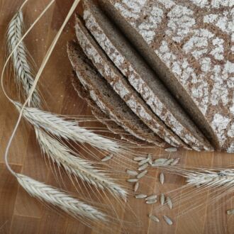 A round loaf of dark rye bread, partly sliced, is on a wooden counter together with dried stalks of rye plants.