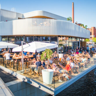 On a sunny day, people sit on a waterside terrace in front of a stylish modern building.