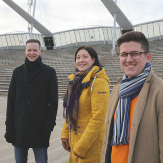 A woman and two men in jackets and scarves stand outside in front of rows of seats at a stadium.
