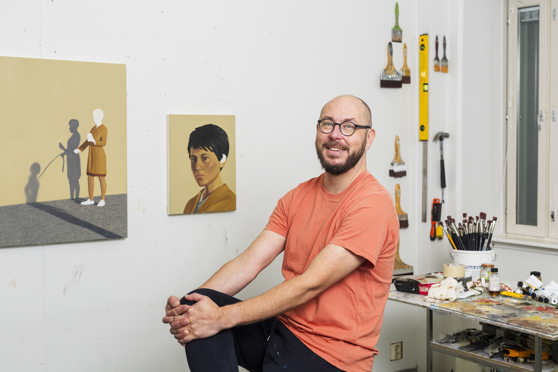 Un homme est assis dans un atelier d’artiste au milieu de pinceaux et d’outils divers, tandis que des tableaux sont visibles au mur.