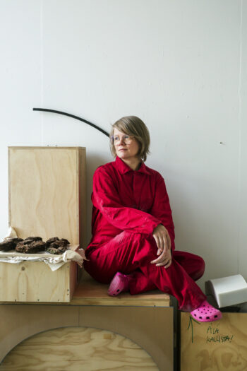 Une femme habillée d’une tenue rouge et de chaussures de type « Crocs » roses est assise sur une boîte en bois.