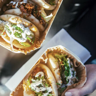 Hands are holding small cardboard trays containing pieces of pita bread with fillings and sauce.