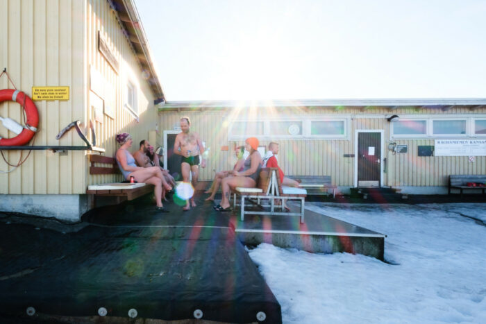 Several people in bathing suits sit on benches beside a wooden building, with snow covering the ground.