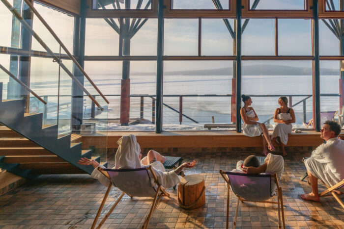 Several people in bathrobes lounge at a spa, with a wintery landscape visible through the windows.