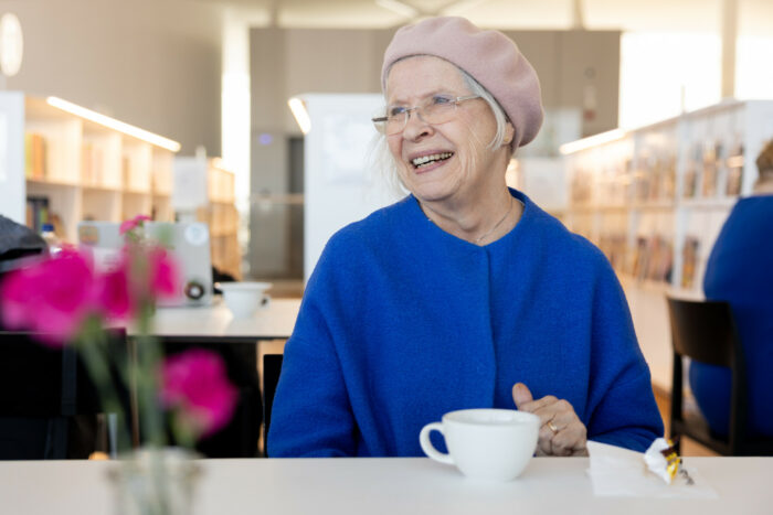 A stylishly dressed older woman sits at a table in front of a cup of coffee and a vase of pink flowers.