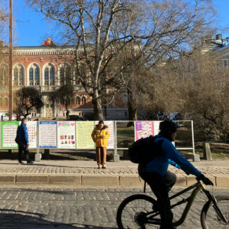 People are looking at a row of posters in metal frames beside the sidewalk in front of a stately building with tall windows.