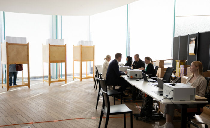 In a large room, people are sitting at a long table, with several voting booths in the background.