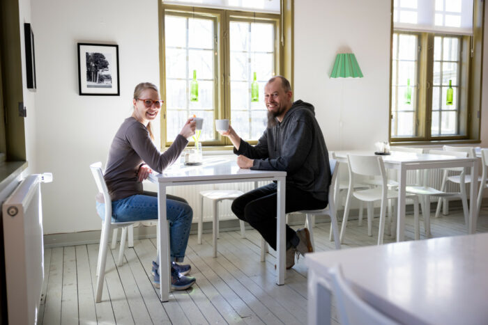 A woman and a man are sitting in a sunlit café and smiling and holding coffee cups.