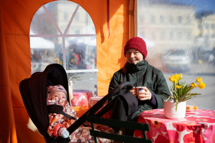 Beside a baby in a stroller, a smiling woman holding a coffee cup sits at a table with a pot of daffodils on it.